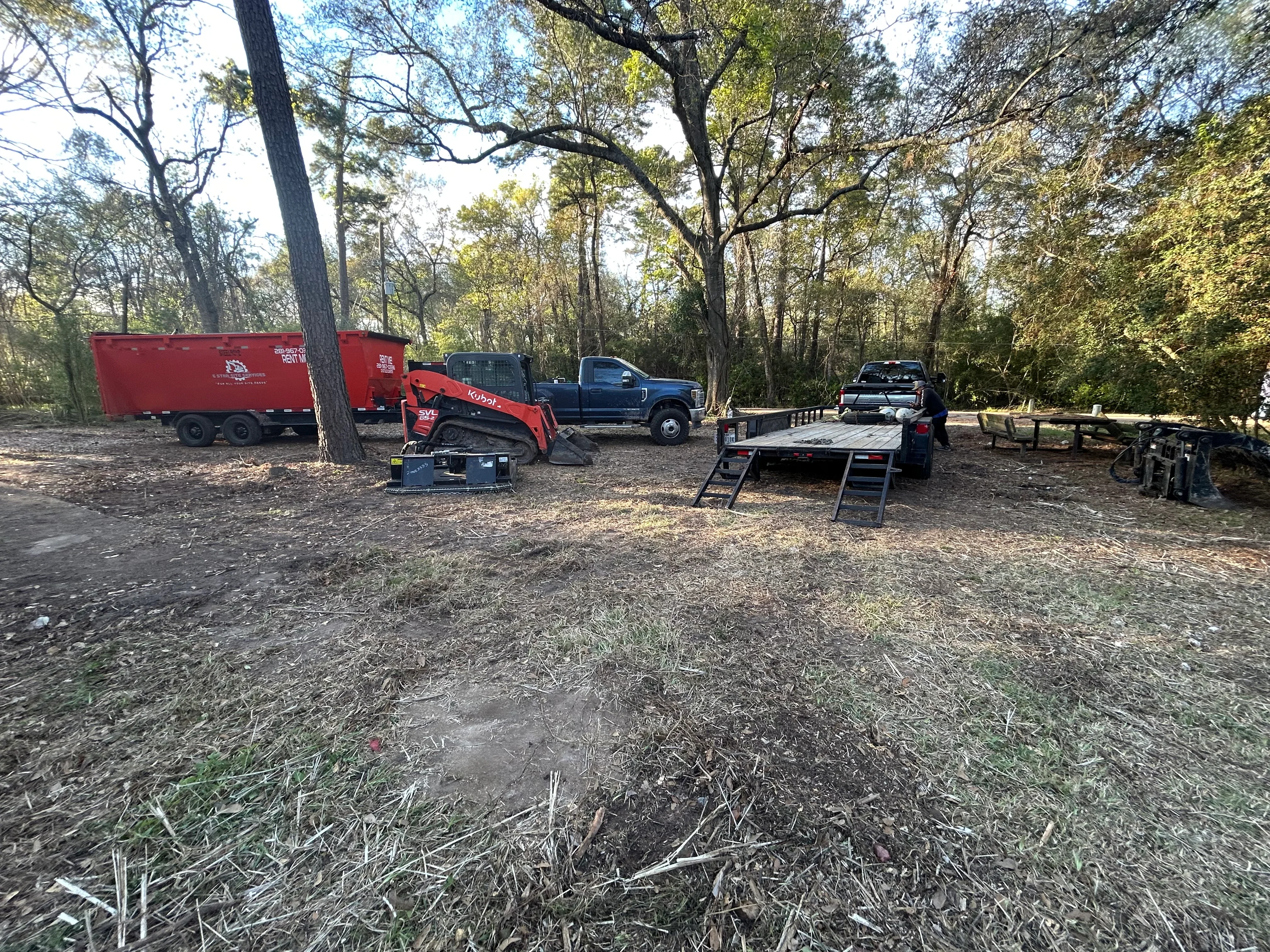 Kubota track loader and dump trailer clearing wooded site with mature trees and debris removal equipment staged