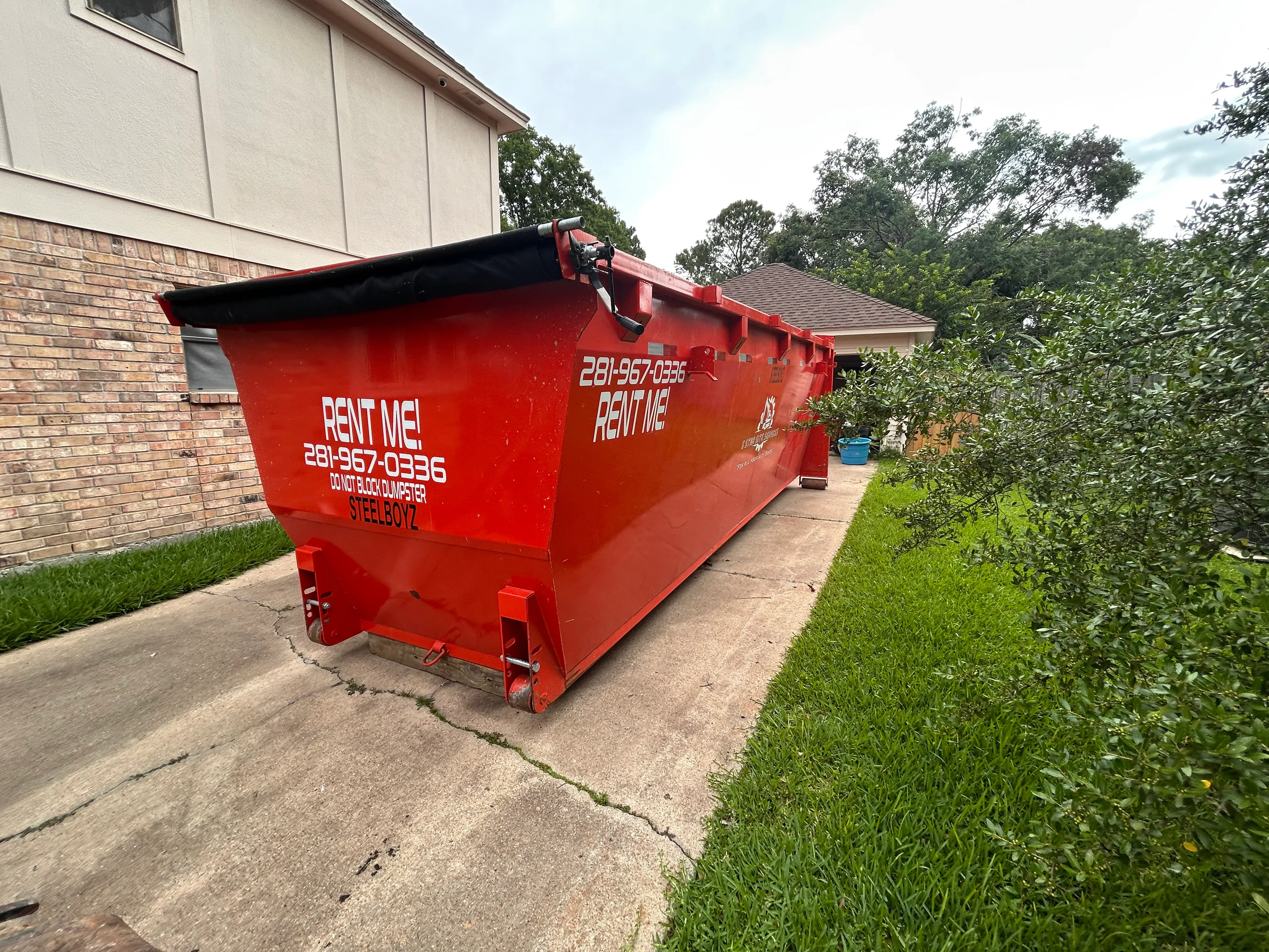 Red steel dumpster bin on residential driveway with hinged cover and wheels